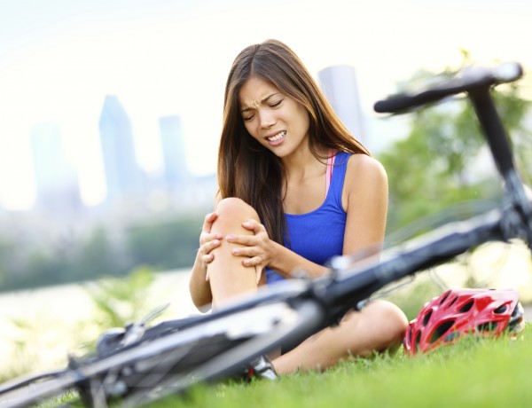 Knee pain bike injury. Woman with pain in knee joints after biking on bicycle. Girl sitting down with painful face expression. Mixed race sport fitness model outdoors.
